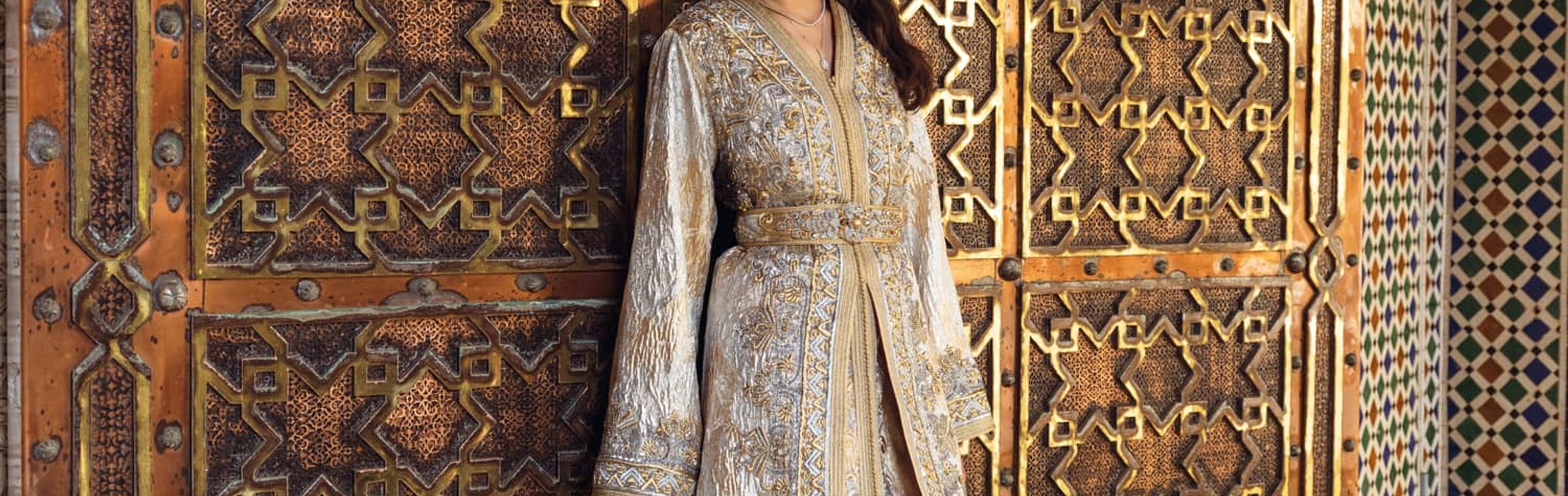 Pearl Gate ivory and silver embroidered Moroccan kaftan with a beaded belt, before an engraved brass door